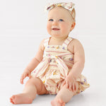 A smiling baby sits on the floor in the Mabel and Honey Purple Bouquet Tank Romper with a matching bow headband, against a plain white background.