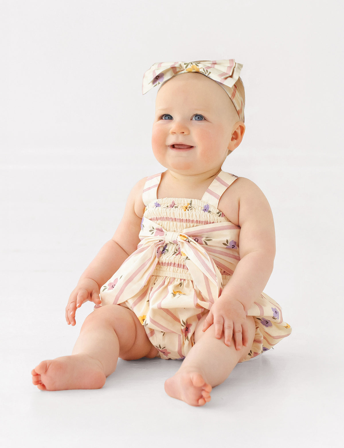 A smiling baby sits on the floor in the Mabel and Honey Purple Bouquet Tank Romper with a matching bow headband, against a plain white background.
