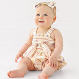 A smiling baby sits on the floor in the Mabel and Honey Purple Bouquet Tank Romper with a matching bow headband, against a plain white background.