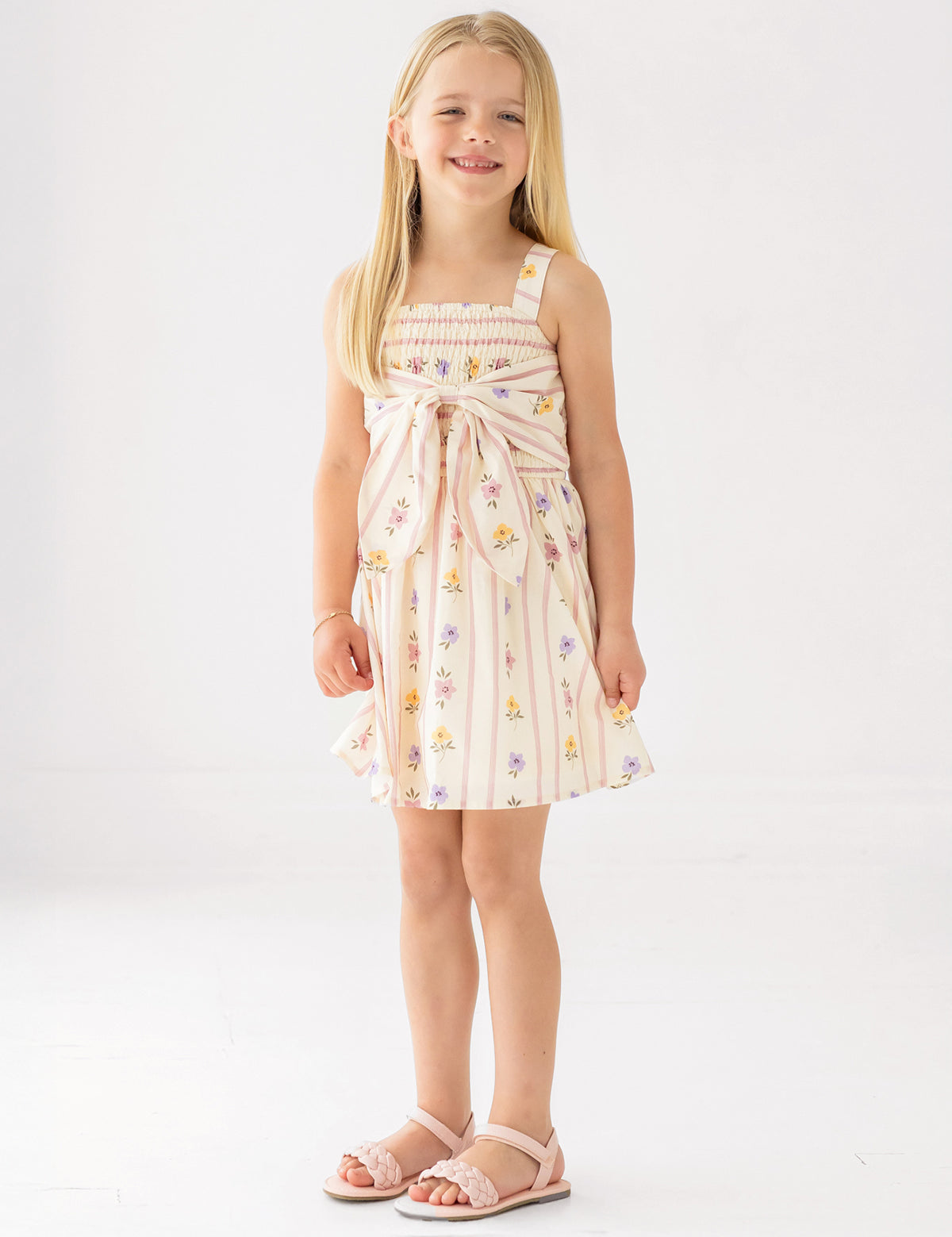 A young girl smiles in the Mabel and Honey Purple Bouquet Tank Dress featuring a smocked bodice and waist bow, paired with pink sandals, against a plain white background.