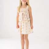 A young girl smiles in the Mabel and Honey Purple Bouquet Tank Dress featuring a smocked bodice and waist bow, paired with pink sandals, against a plain white background.