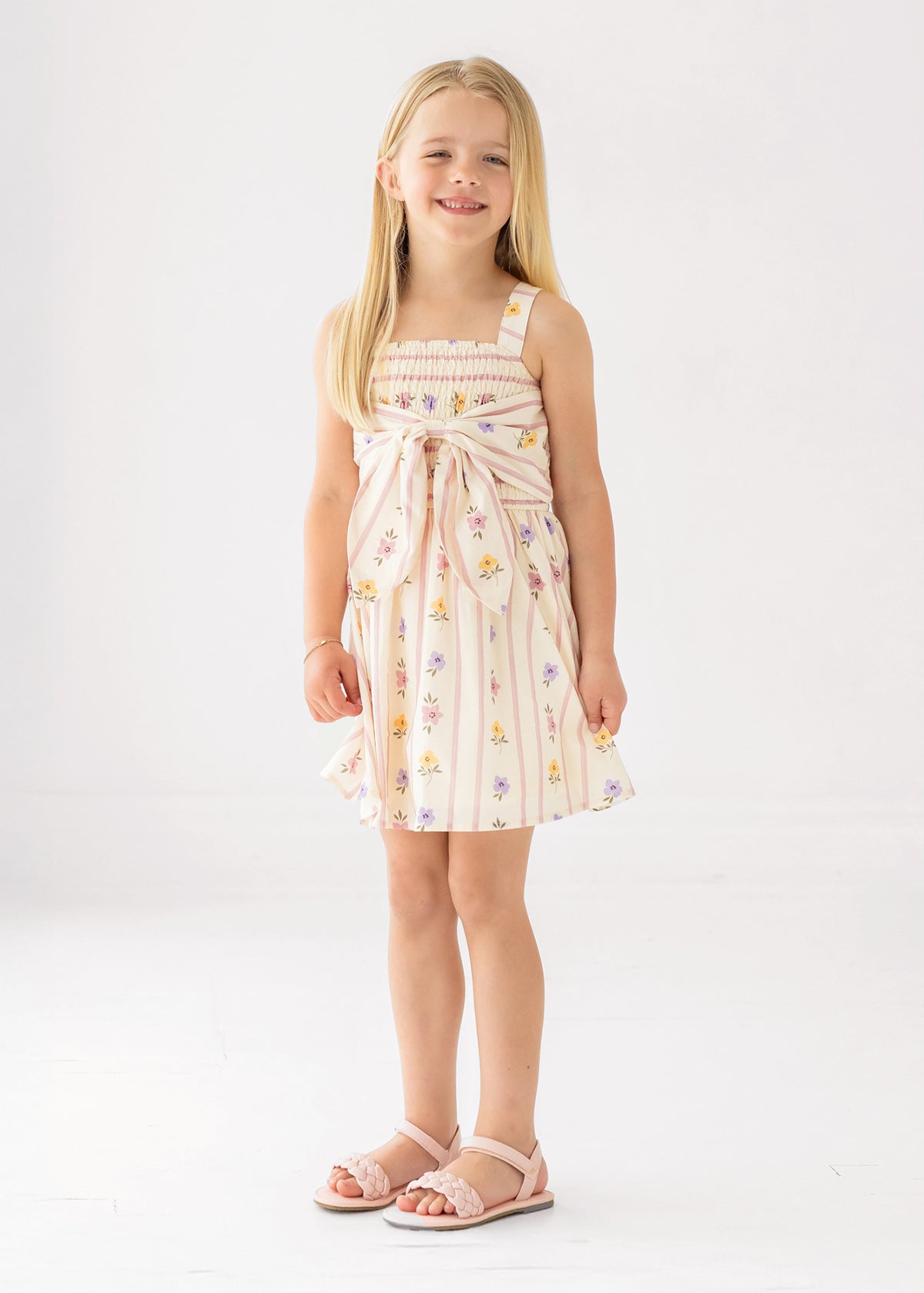 A young girl smiles in the Mabel and Honey Purple Bouquet Tank Dress featuring a smocked bodice and waist bow, paired with pink sandals, against a plain white background.