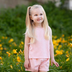 A young girl with long blonde pigtails smiles outdoors in front of green bushes and yellow flowers, wearing the Mabel and Honey Pink Plaid Perfection Sleeveless Two Piece Set.
