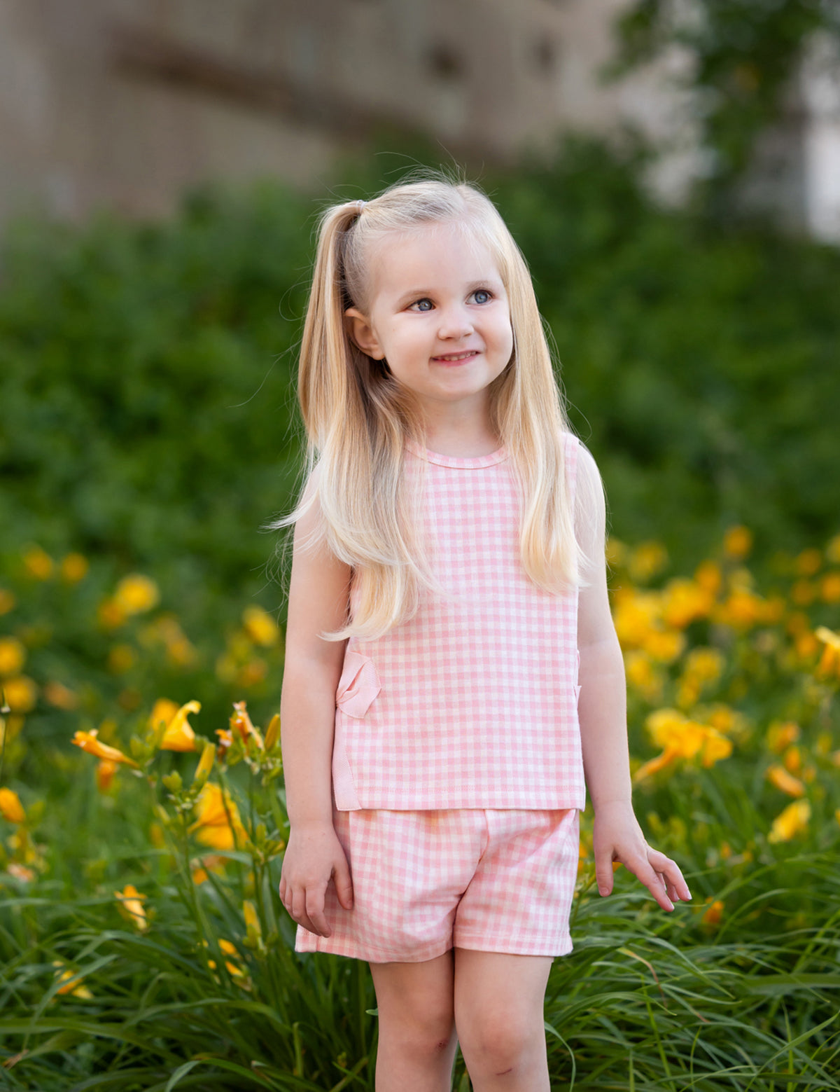 A young girl with long blonde pigtails smiles outdoors in front of green bushes and yellow flowers, wearing the Mabel and Honey Pink Plaid Perfection Sleeveless Two Piece Set.