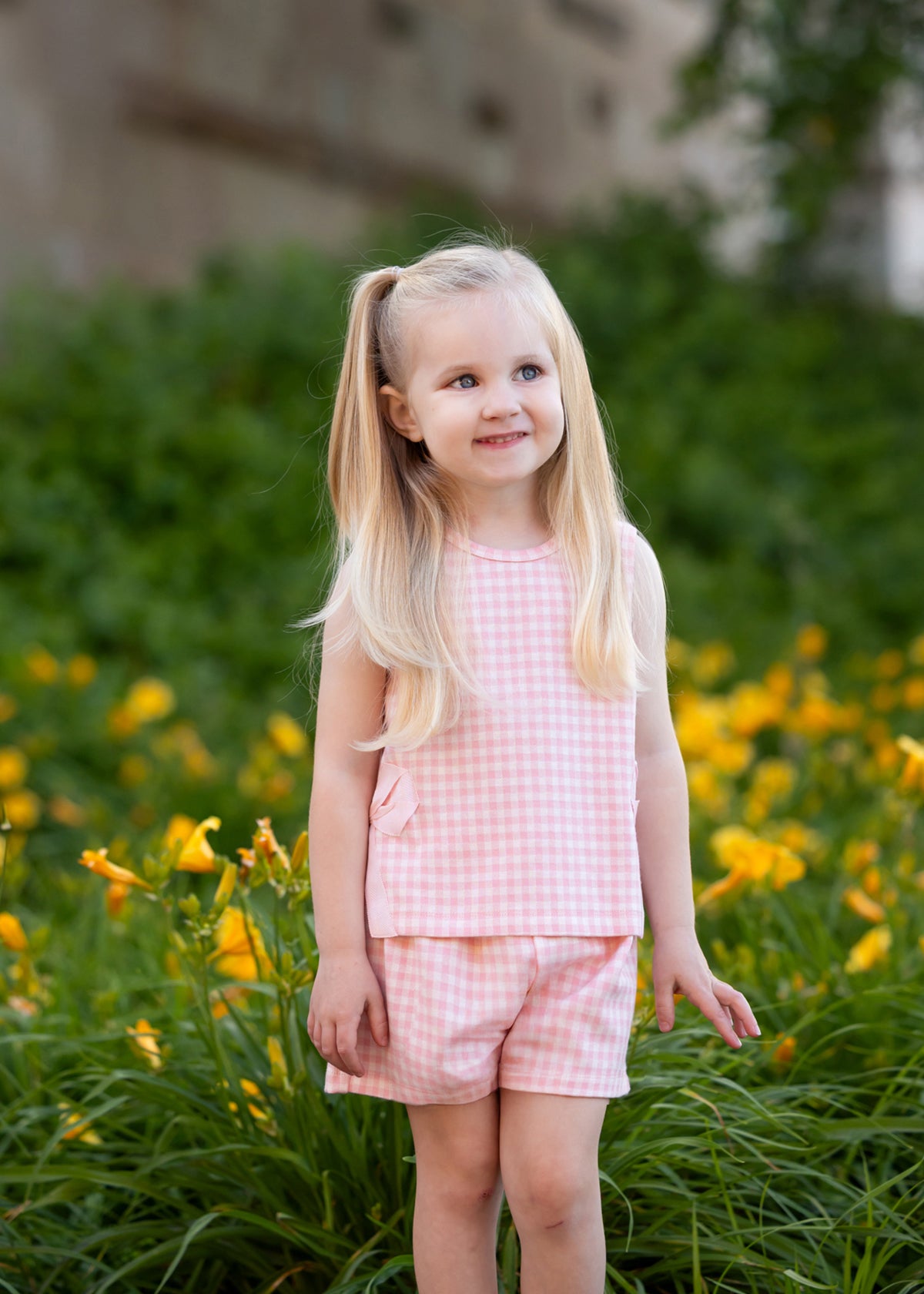 A young girl with long blonde pigtails smiles outdoors in front of green bushes and yellow flowers, wearing the Mabel and Honey Pink Plaid Perfection Sleeveless Two Piece Set.