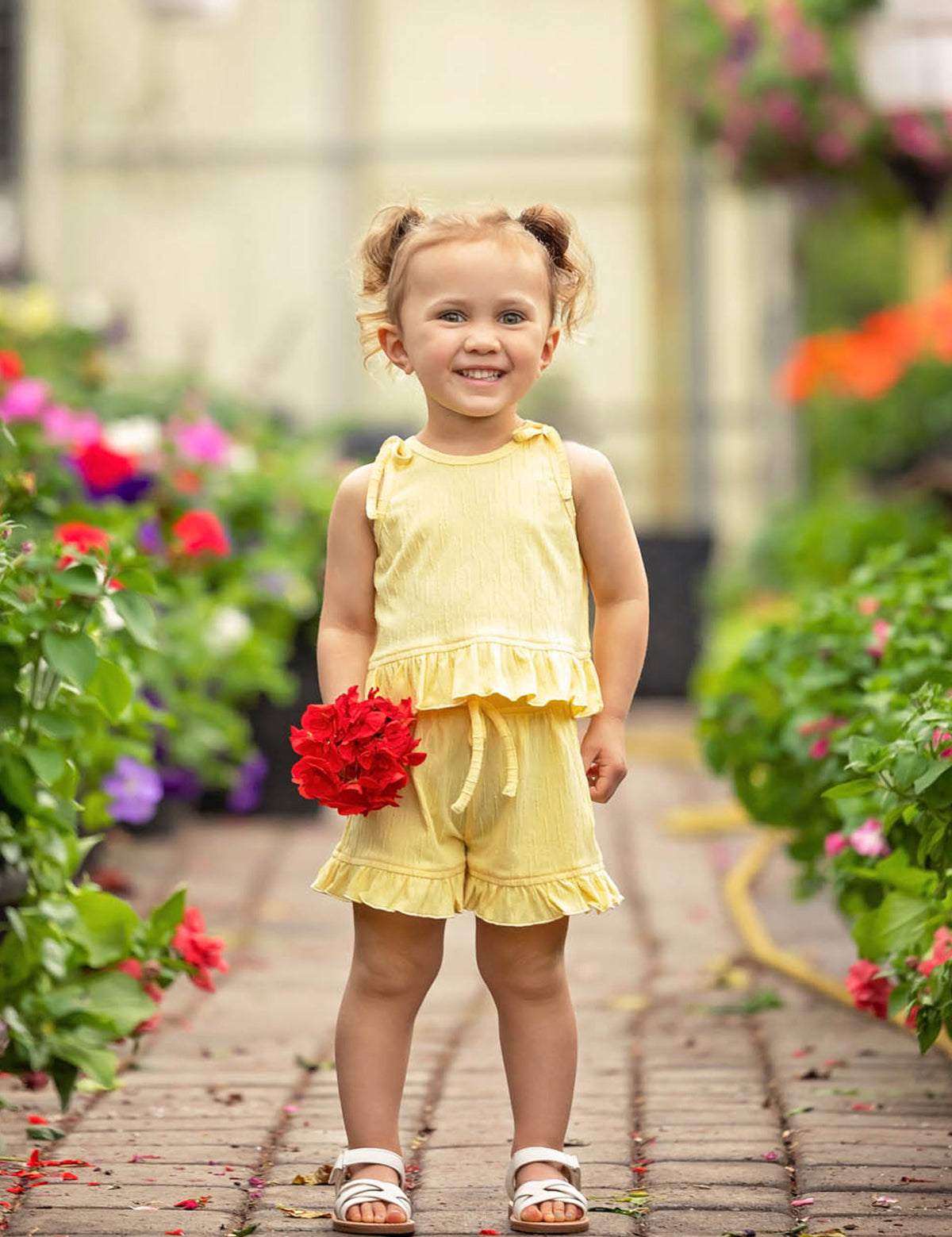 A smiling young girl with light brown hair in pigtails stands on a garden path, surrounded by colorful blossoms, wearing the Sonny Sleeveless with Knot Detail Two Piece Set from Mabel and Honey and white sandals, holding a bunch of red flowers.