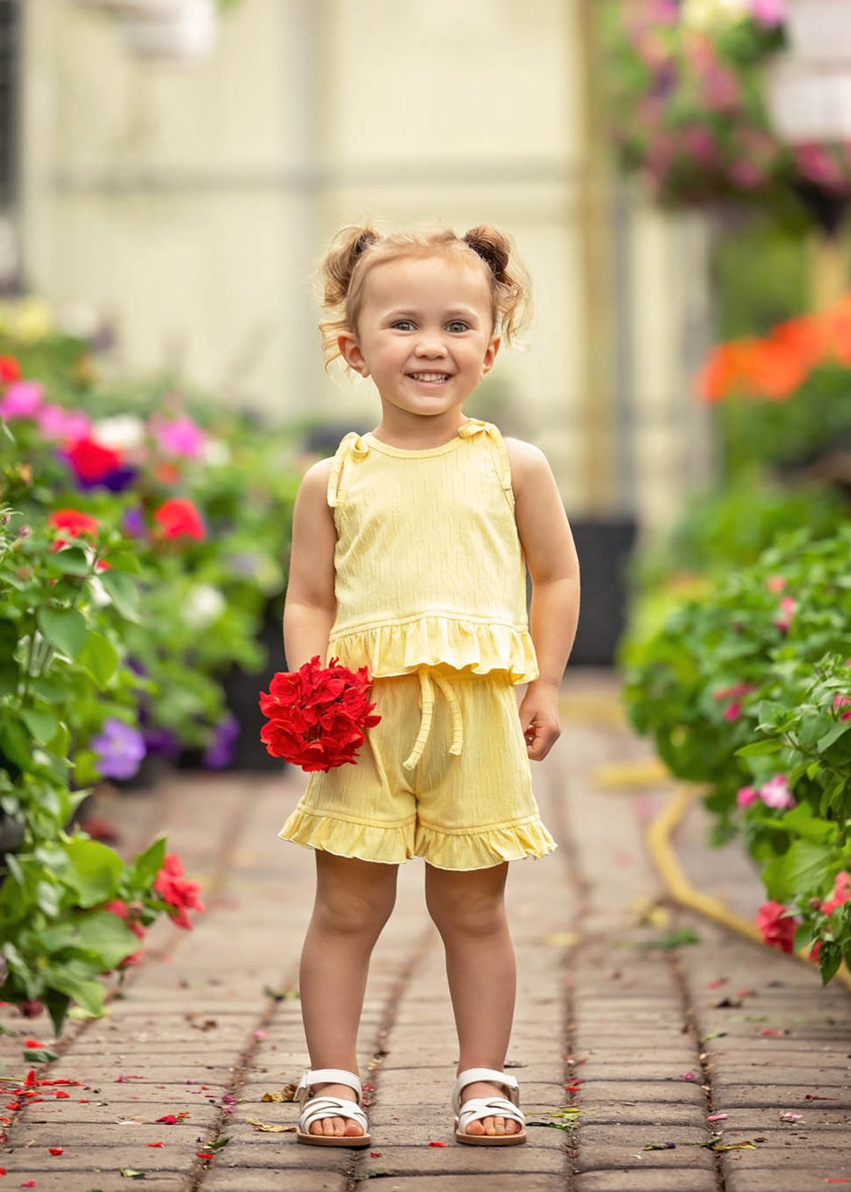 A smiling young girl with light brown hair in pigtails stands on a garden path, surrounded by colorful blossoms, wearing the Sonny Sleeveless with Knot Detail Two Piece Set from Mabel and Honey and white sandals, holding a bunch of red flowers.