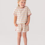 A young girl smiles with her hands in the pockets of her Mabel and Honey Sunset Sherbet Short Sleeve Two Piece Set—featuring a striped top and matching shorts—paired with white sneakers against a plain, light background.