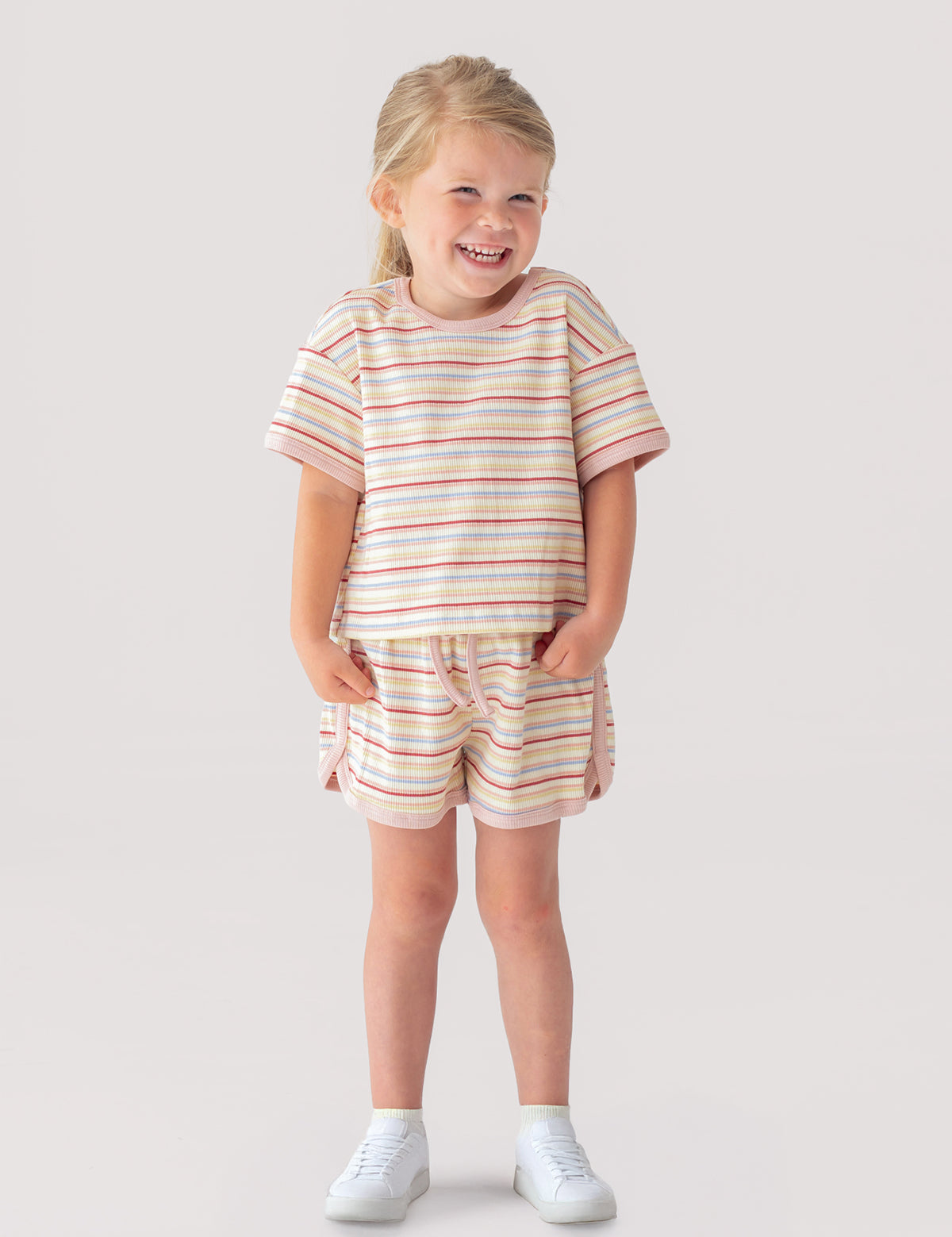 A young girl smiles with her hands in the pockets of her Mabel and Honey Sunset Sherbet Short Sleeve Two Piece Set—featuring a striped top and matching shorts—paired with white sneakers against a plain, light background.