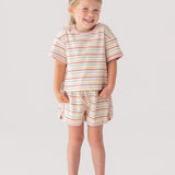 A young girl smiles with her hands in the pockets of her Mabel and Honey Sunset Sherbet Short Sleeve Two Piece Set—featuring a striped top and matching shorts—paired with white sneakers against a plain, light background.