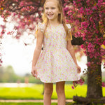 A young girl smiles in the sun, standing on grass before a tree with bright pink blossoms, wearing the Sweet Melody Short Flutter Sleeve Dress by Mabel and Honey.