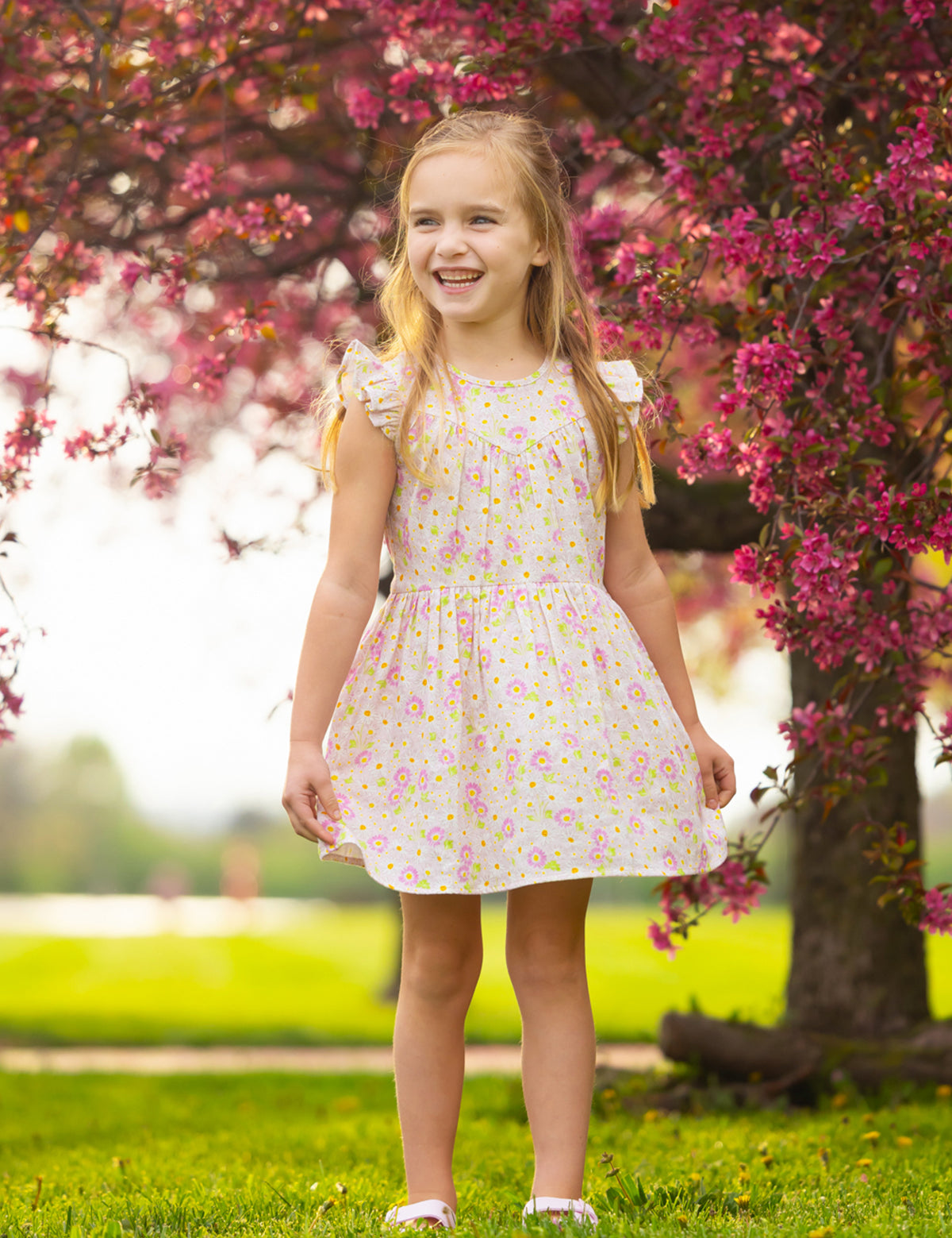 A young girl smiles in the sun, standing on grass before a tree with bright pink blossoms, wearing the Sweet Melody Short Flutter Sleeve Dress by Mabel and Honey.