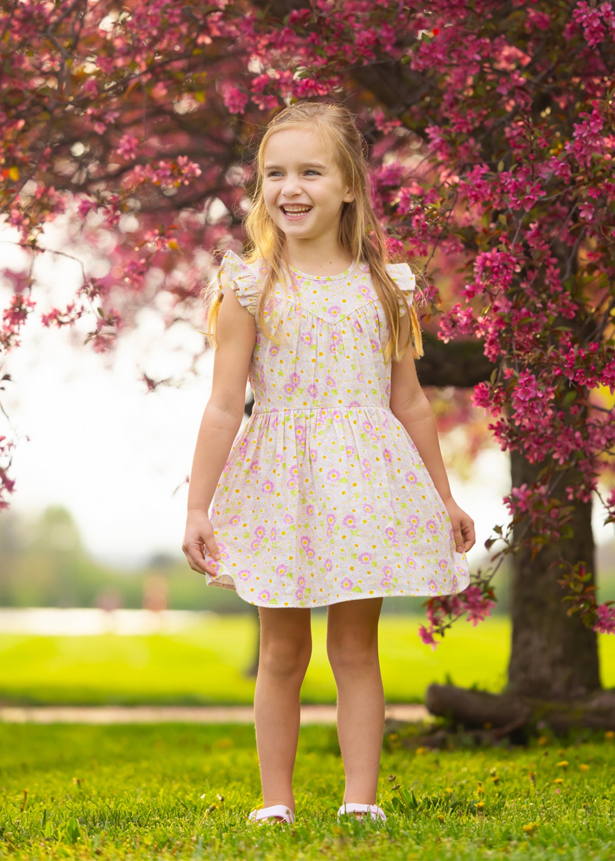 A young girl smiles in the sun, standing on grass before a tree with bright pink blossoms, wearing the Sweet Melody Short Flutter Sleeve Dress by Mabel and Honey.