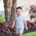 A young boy wears the Beckett and Bear Eli Short Sleeve Two Piece Set, walking barefoot and smiling on green grass with trees and red-leafed plants in the background.