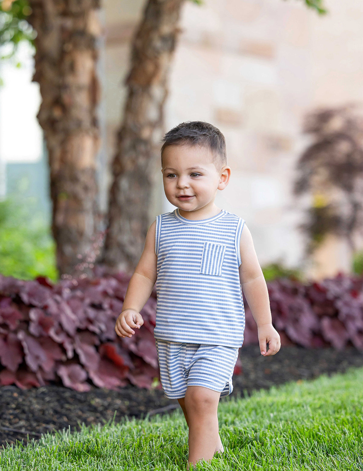 A young boy wears the Beckett and Bear Eli Short Sleeve Two Piece Set, walking barefoot and smiling on green grass with trees and red-leafed plants in the background.