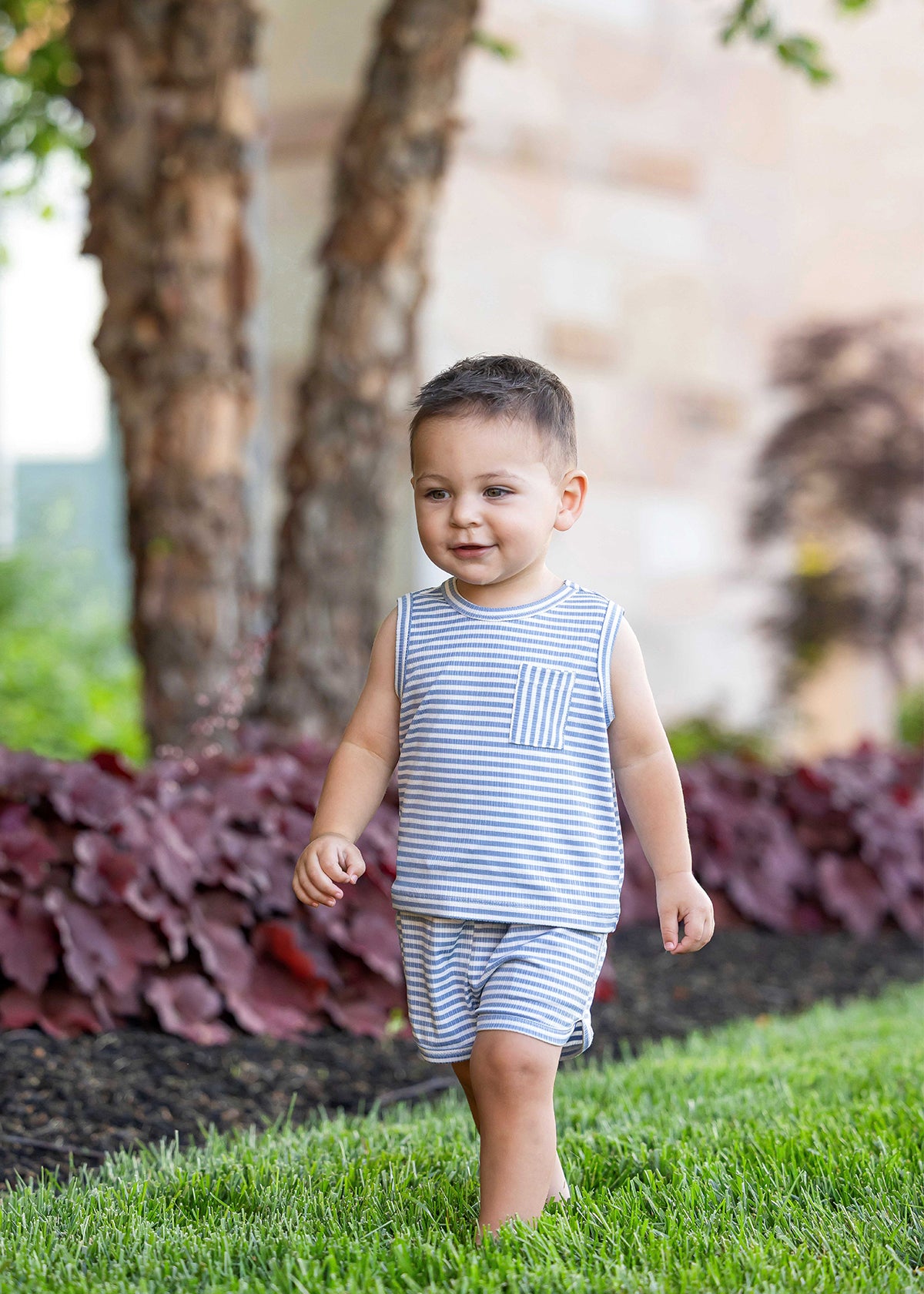 A young boy wears the Beckett and Bear Eli Short Sleeve Two Piece Set, walking barefoot and smiling on green grass with trees and red-leafed plants in the background.