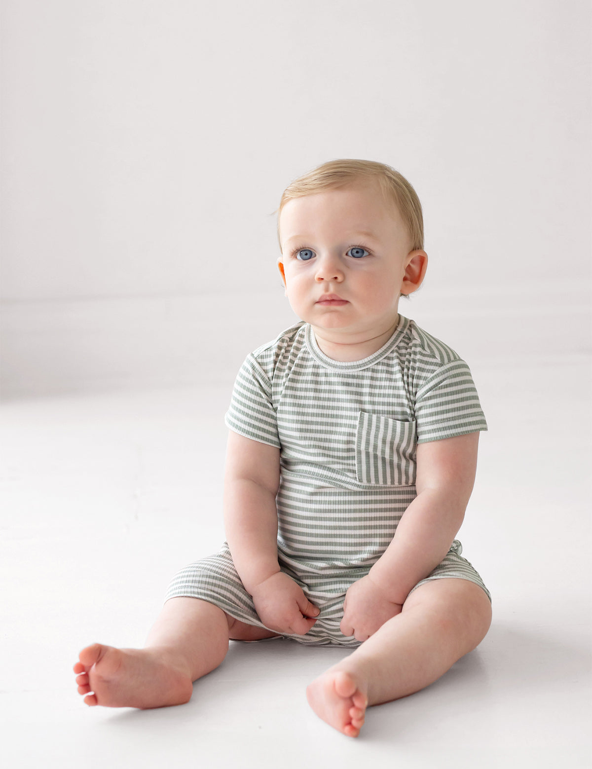 A light-skinned, blue-eyed baby sits calmly on a white floor, wearing the Beckett and Bear Frankie Short Sleeve Romper in sage green with white stripes and chest pocket. The stylish one-piece adds charm as the baby looks slightly to the side.