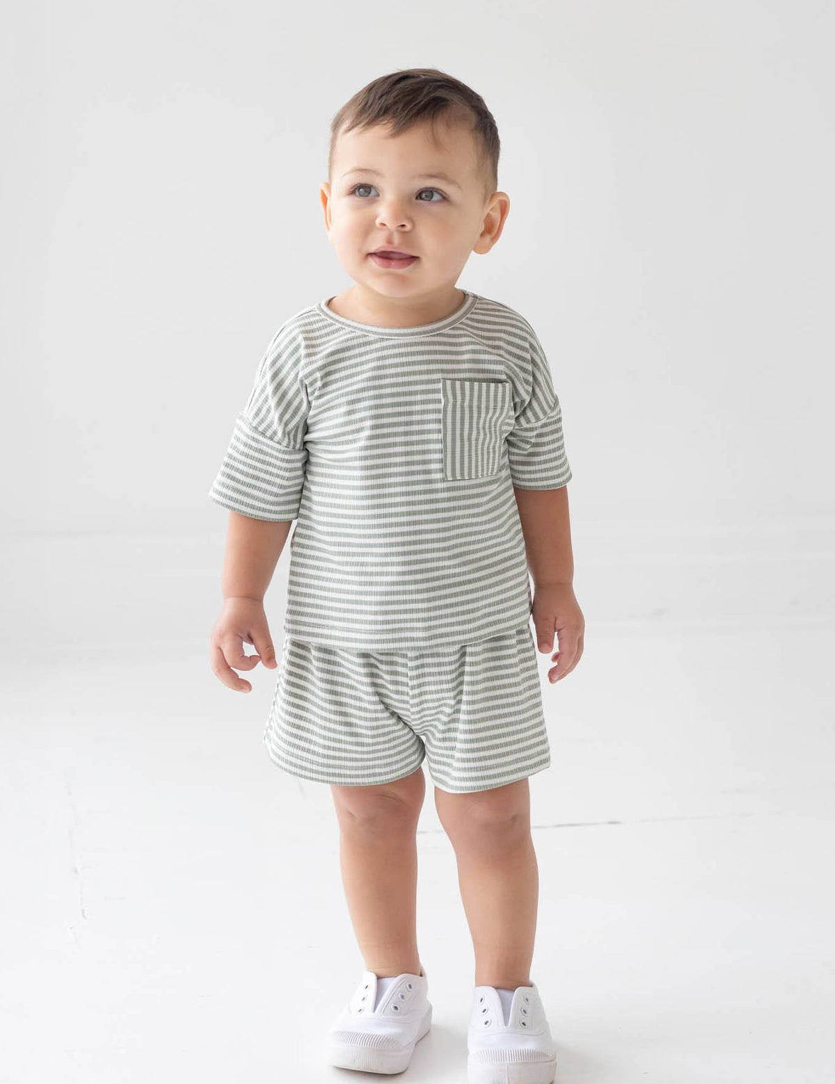 A toddler stands indoors wearing the Beckett and Bear Frankie Short Sleeve Two Piece Set, a matching gray and white striped outfit with shorts and a short-sleeve shirt, paired with white sneakers. He has short brown hair and a soft smile as he looks slightly upward.