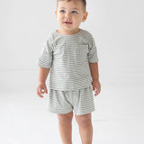 A toddler stands indoors wearing the Beckett and Bear Frankie Short Sleeve Two Piece Set, a matching gray and white striped outfit with shorts and a short-sleeve shirt, paired with white sneakers. He has short brown hair and a soft smile as he looks slightly upward.