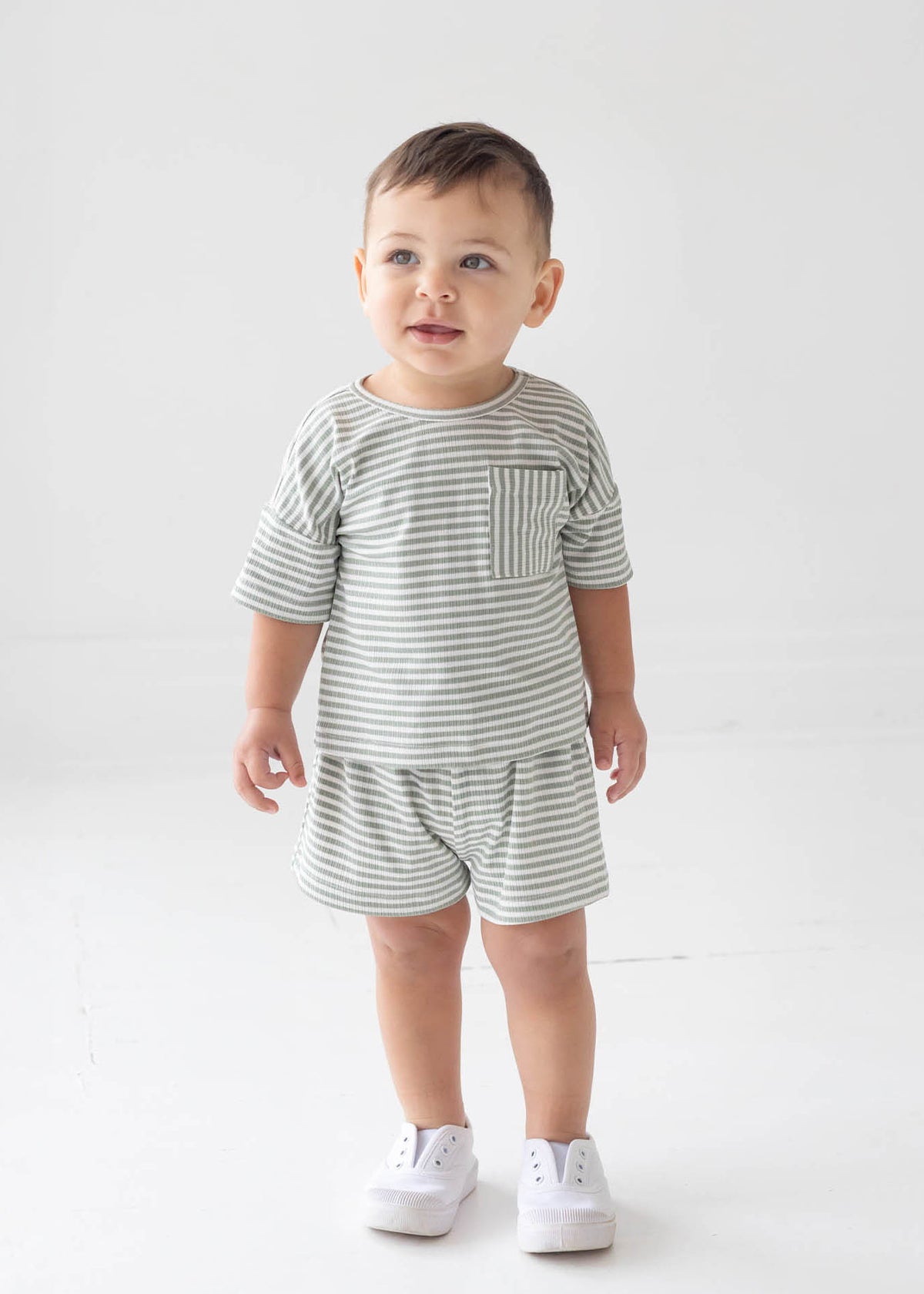 A toddler stands indoors wearing the Beckett and Bear Frankie Short Sleeve Two Piece Set, a matching gray and white striped outfit with shorts and a short-sleeve shirt, paired with white sneakers. He has short brown hair and a soft smile as he looks slightly upward.