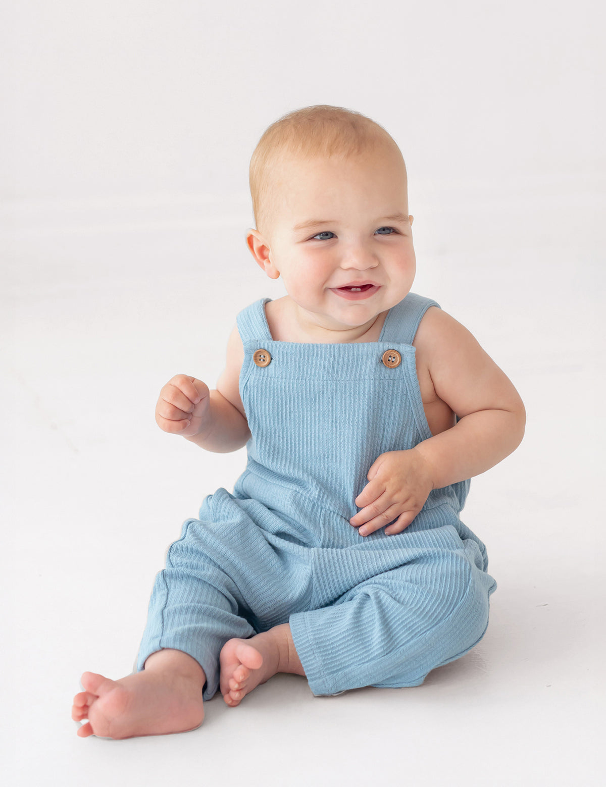 A smiling baby in the Caleb Tank Romper by Beckett and Bear sits barefoot on a white surface, looking slightly to the side. The light blue, ribbed fabric of this cozy one-piece adds a charming touch to the adorable scene.