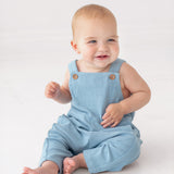 A smiling baby in the Caleb Tank Romper by Beckett and Bear sits barefoot on a white surface, looking slightly to the side. The light blue, ribbed fabric of this cozy one-piece adds a charming touch to the adorable scene.