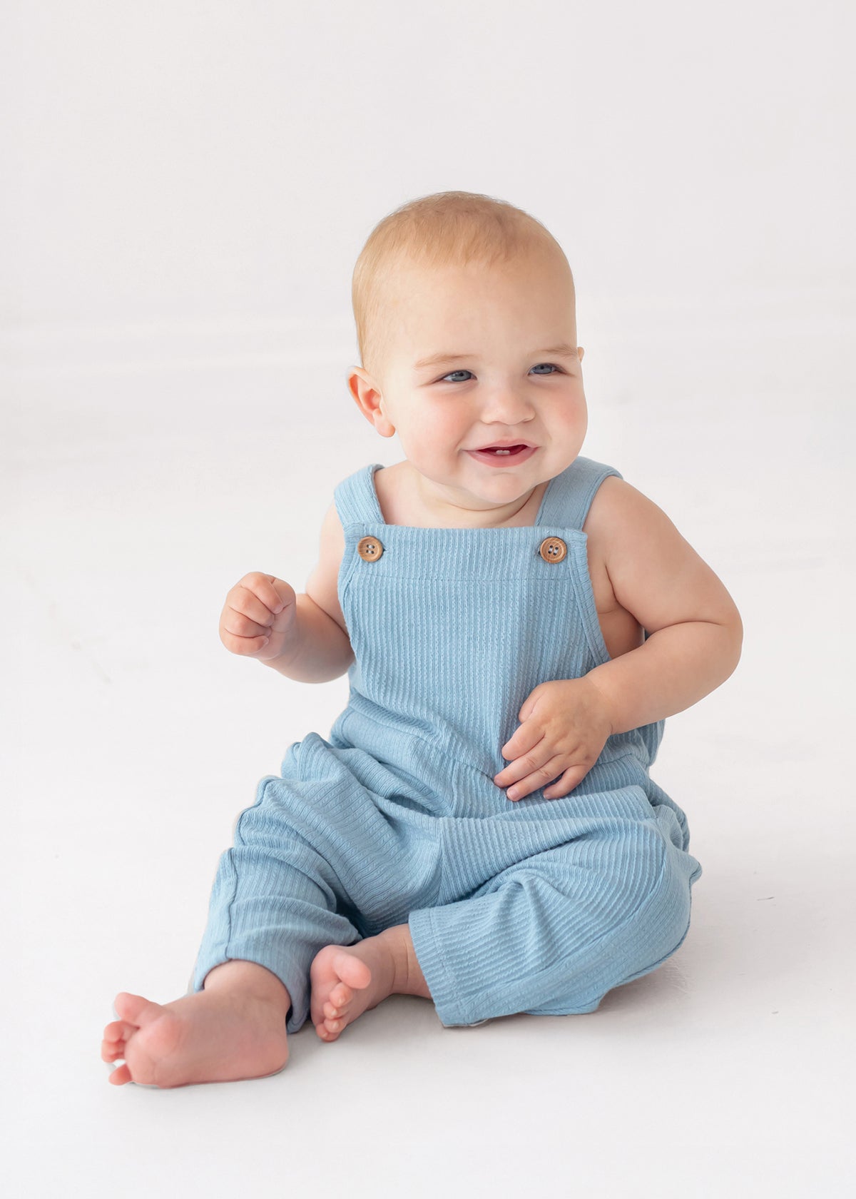 A smiling baby in the Caleb Tank Romper by Beckett and Bear sits barefoot on a white surface, looking slightly to the side. The light blue, ribbed fabric of this cozy one-piece adds a charming touch to the adorable scene.
