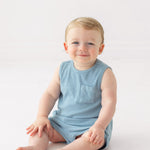 A smiling baby with light hair sits on the floor wearing the Beckett and Bear Caleb Sleeveless Two Piece Set, against a plain white background.