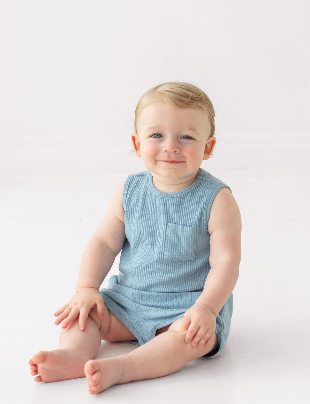 A smiling baby with light hair sits on the floor wearing the Beckett and Bear Caleb Sleeveless Two Piece Set, against a plain white background.