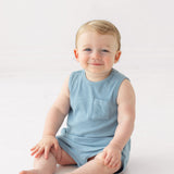 A smiling baby with light hair sits on the floor wearing the Beckett and Bear Caleb Sleeveless Two Piece Set, against a plain white background.