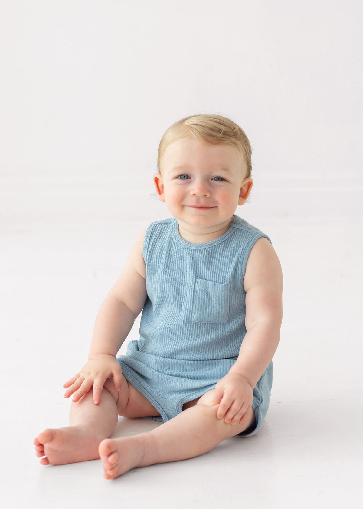 A smiling baby with light hair sits on the floor wearing the Beckett and Bear Caleb Sleeveless Two Piece Set, against a plain white background.