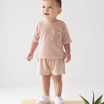 A toddler stands indoors on a wooden block, wearing the Beckett and Bear Bear Short Sleeve Two Piece Set in light pink with white slip-on shoes. A green potted plant sits in the lower right corner against a white background.