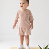 A toddler stands indoors on a wooden block, wearing the Beckett and Bear Bear Short Sleeve Two Piece Set in light pink with white slip-on shoes. A green potted plant sits in the lower right corner against a white background.