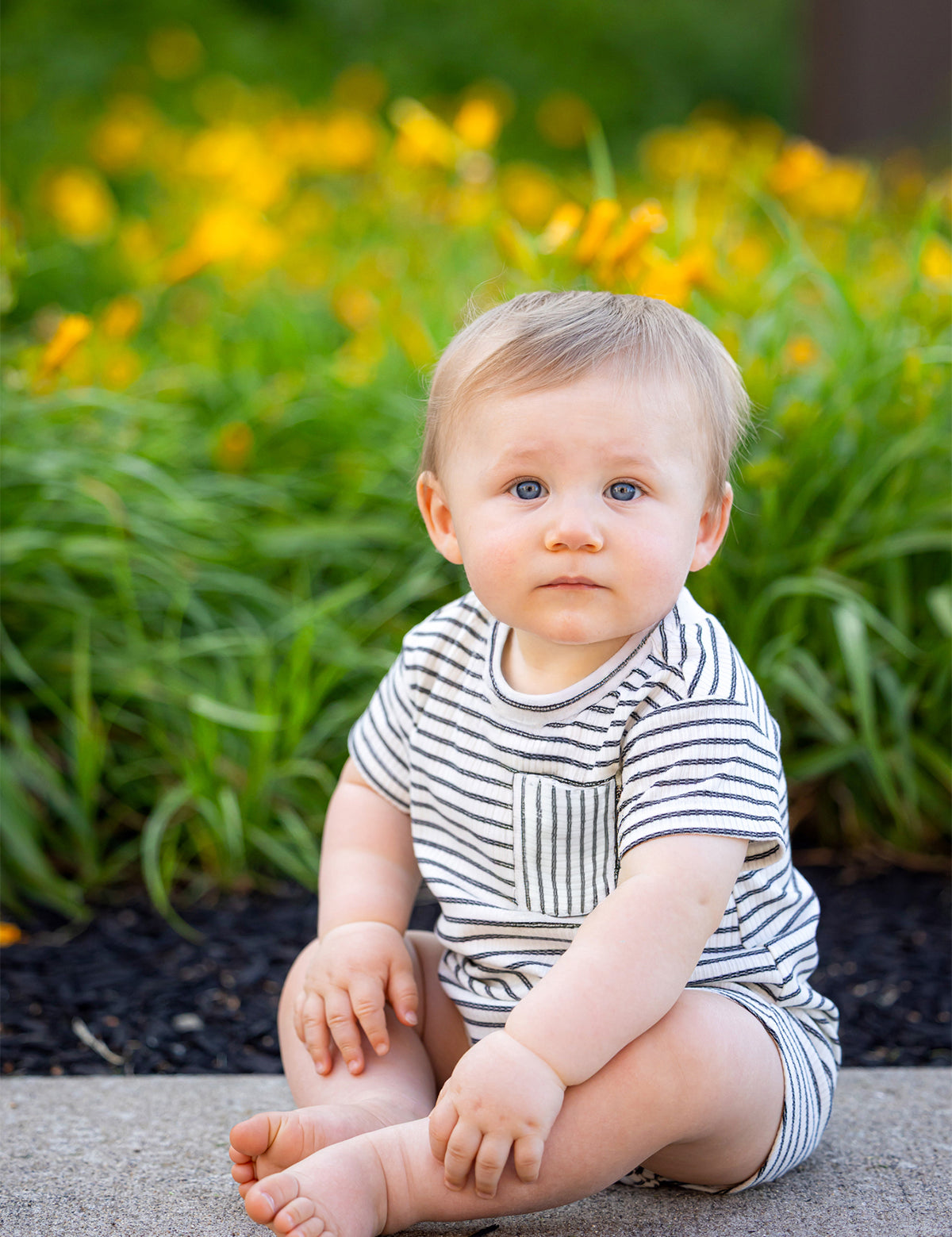 A baby with blond hair and blue eyes sits on a sidewalk by green plants and yellow flowers, wearing the Beckett and Bear Micah Short Sleeve Romper, and looks at the camera.