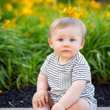 A baby with blond hair and blue eyes sits on a sidewalk by green plants and yellow flowers, wearing the Beckett and Bear Micah Short Sleeve Romper, and looks at the camera.