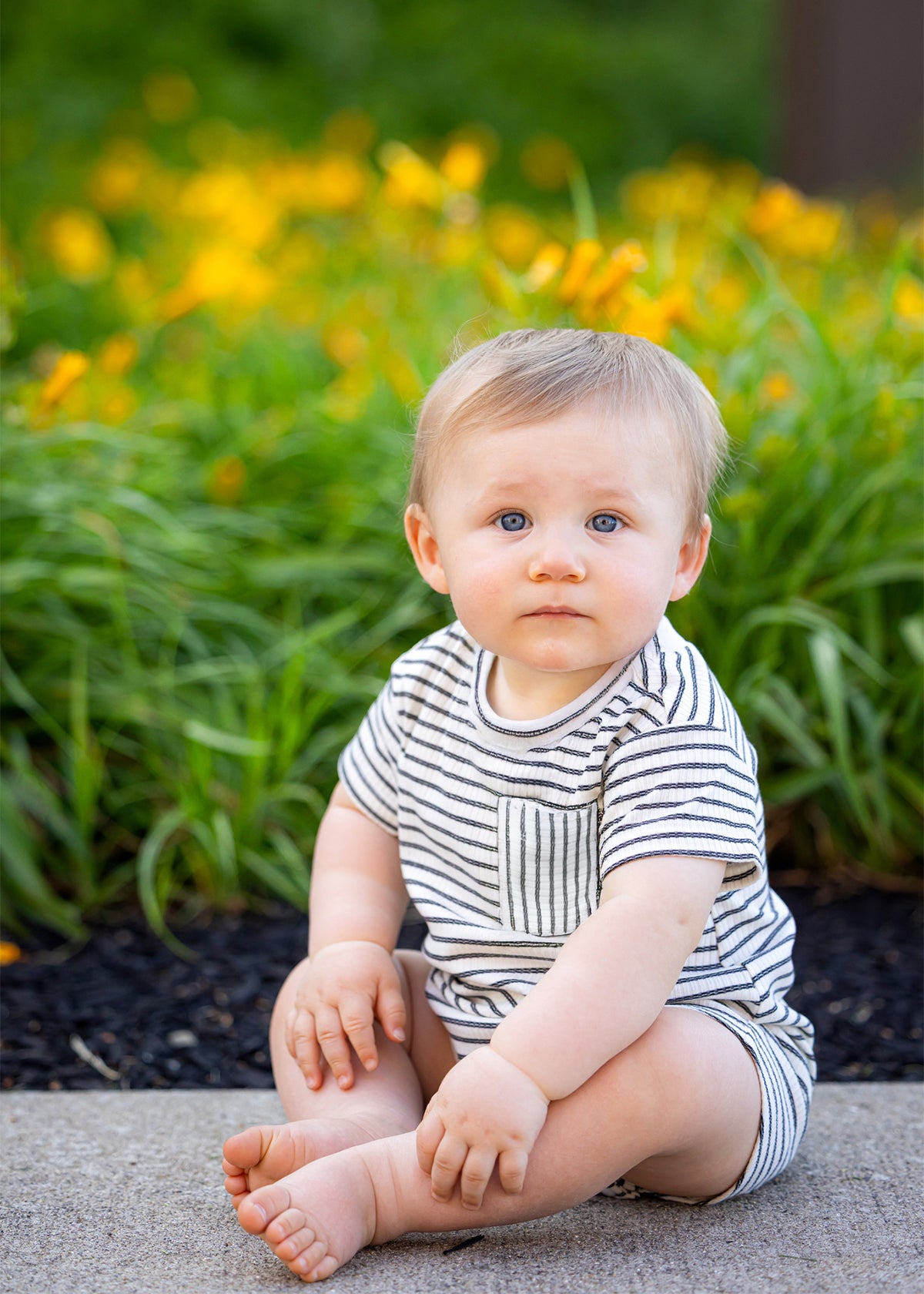 A baby with blond hair and blue eyes sits on a sidewalk by green plants and yellow flowers, wearing the Beckett and Bear Micah Short Sleeve Romper, and looks at the camera.