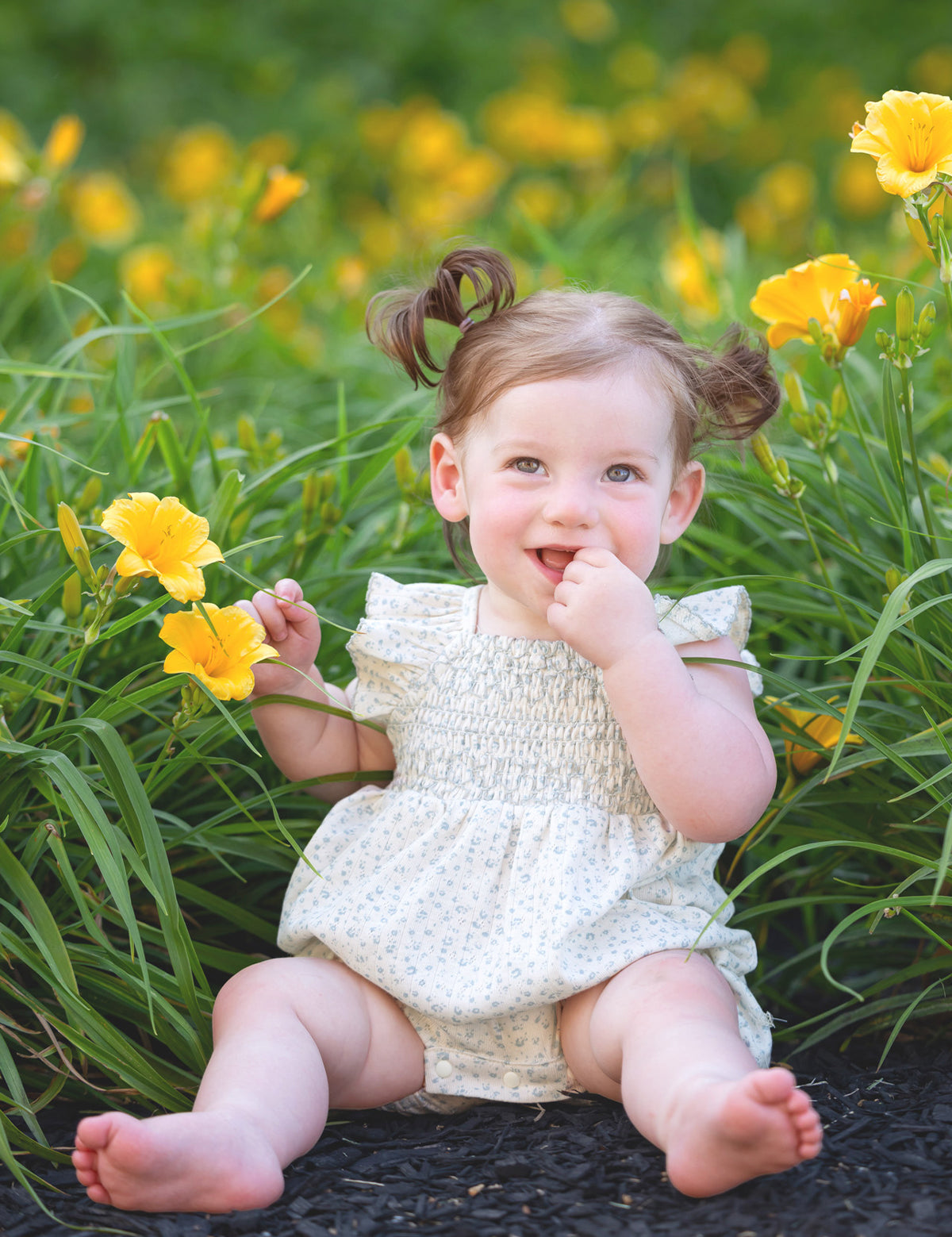 A smiling toddler with light brown hair in pigtails sits among yellow flowers and green grass, playfully touching her mouth while wearing the Mabel and Honey Opal Short Flutter Sleeve Romper.