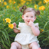 A smiling toddler with light brown hair in pigtails sits among yellow flowers and green grass, playfully touching her mouth while wearing the Mabel and Honey Opal Short Flutter Sleeve Romper.