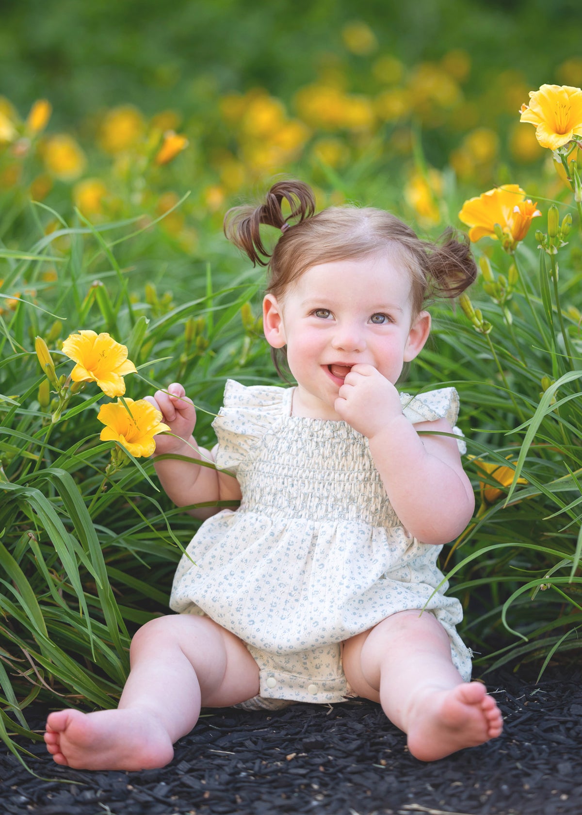 A smiling toddler with light brown hair in pigtails sits among yellow flowers and green grass, playfully touching her mouth while wearing the Mabel and Honey Opal Short Flutter Sleeve Romper.