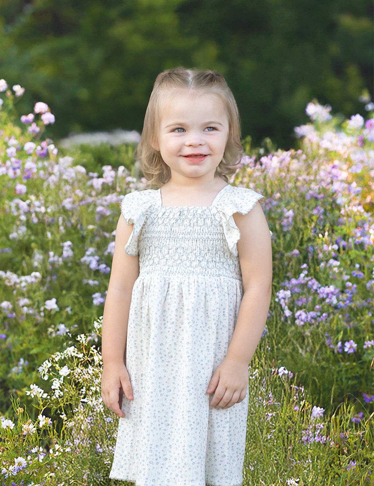 A young child with light hair smiles in a field of purple and white wildflowers, wearing the Opal Short Flutter Sleeve Dress by Mabel and Honey against a backdrop of greenery.