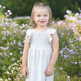 A young child with light hair smiles in a field of purple and white wildflowers, wearing the Opal Short Flutter Sleeve Dress by Mabel and Honey against a backdrop of greenery.