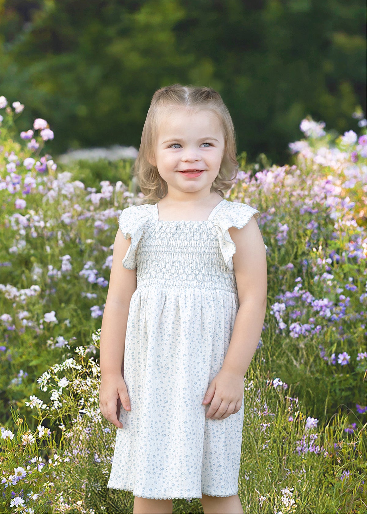 A young child with light hair smiles in a field of purple and white wildflowers, wearing the Opal Short Flutter Sleeve Dress by Mabel and Honey against a backdrop of greenery.
