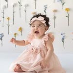 A smiling baby sits on the floor wearing the Isobella and Chloe Sweet Peony Pink Flutter Sleeve Dress and matching headband. Behind her, colorful flowers are attached vertically to a white wall.