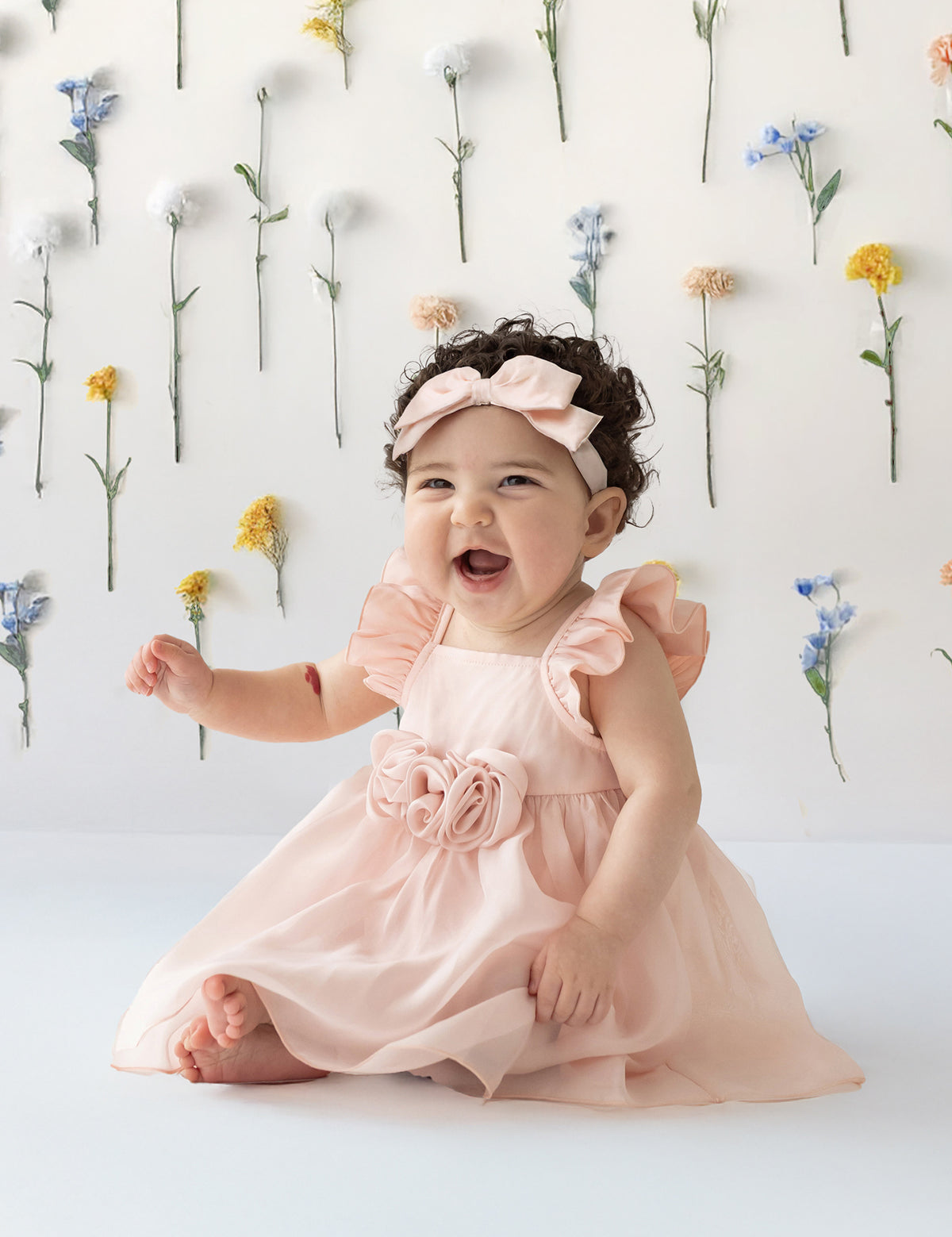 A smiling baby sits on the floor wearing the Isobella and Chloe Sweet Peony Pink Flutter Sleeve Dress and matching headband. Behind her, colorful flowers are attached vertically to a white wall.
