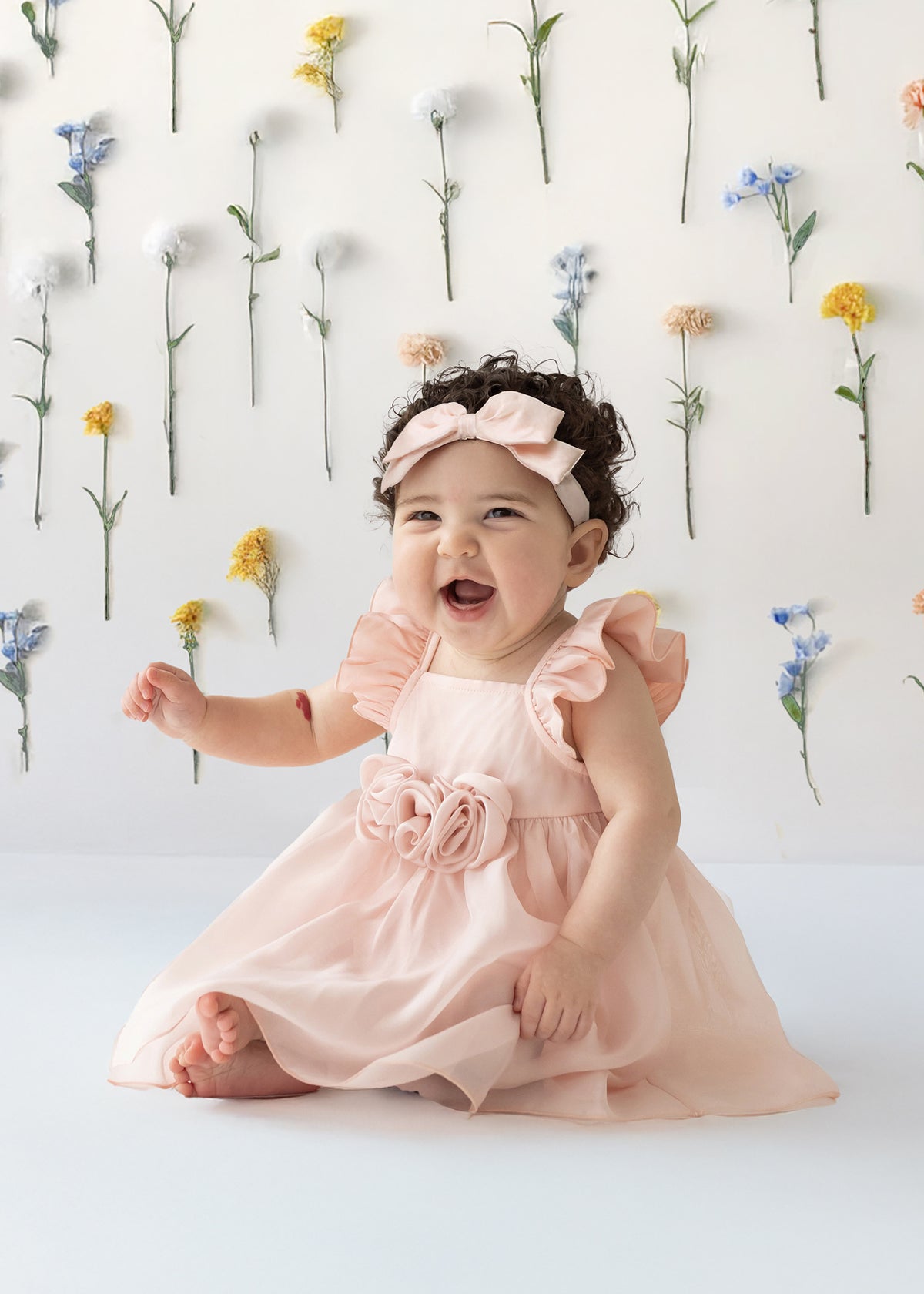 A smiling baby sits on the floor wearing the Isobella and Chloe Sweet Peony Pink Flutter Sleeve Dress and matching headband. Behind her, colorful flowers are attached vertically to a white wall.