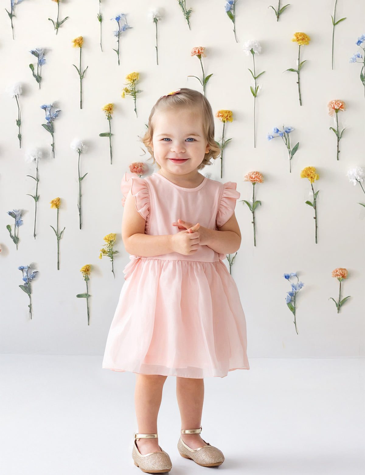 A young girl with light brown hair tied up smiles in front of a white wall adorned with colorful flowers, wearing the Sweet Peony Sleeveless Flutter Sleeve Dress by Isobella and Chloe and beige shoes.
