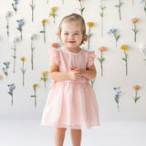 A young girl with light brown hair tied up smiles in front of a white wall adorned with colorful flowers, wearing the Sweet Peony Sleeveless Flutter Sleeve Dress by Isobella and Chloe and beige shoes.