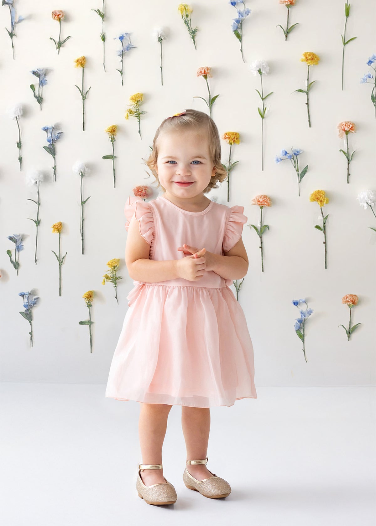 A young girl with light brown hair tied up smiles in front of a white wall adorned with colorful flowers, wearing the Sweet Peony Sleeveless Flutter Sleeve Dress by Isobella and Chloe and beige shoes.