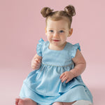 A light-skinned, blue-eyed baby with brown hair in two buns sits barefoot on a pale pink floor, wearing the Pearly Girly Flutter Sleeve Dress by Isobella and Chloe and smiling softly.
