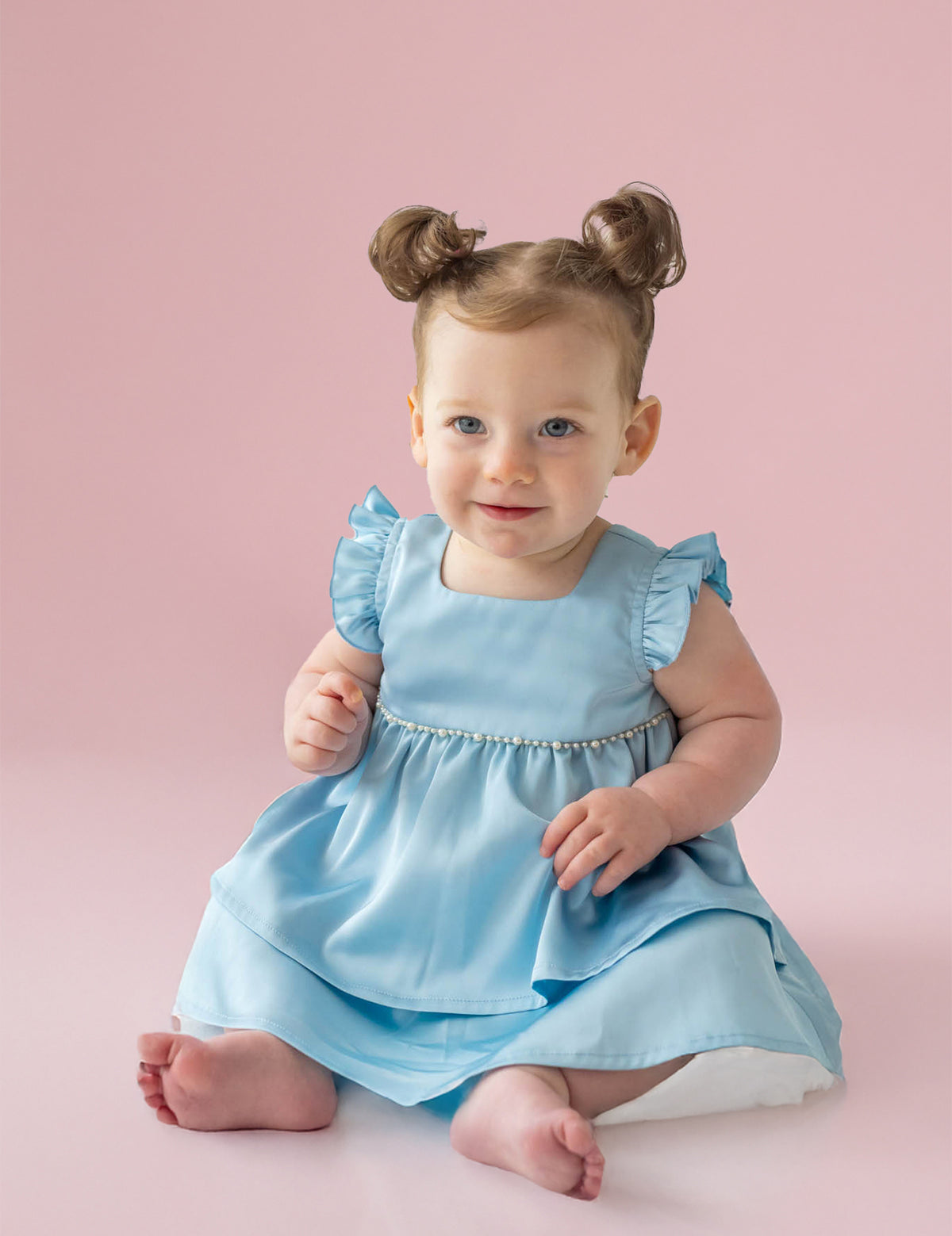 A light-skinned, blue-eyed baby with brown hair in two buns sits barefoot on a pale pink floor, wearing the Pearly Girly Flutter Sleeve Dress by Isobella and Chloe and smiling softly.